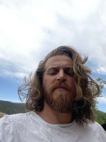 Un homme aux longs cheveux bruns ondulés et à la barbe fournie se tient à l'extérieur, vêtu d'une chemise blanche. Il a l'air détendu, les yeux fermés, sous un ciel nuageux.