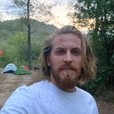 La imagen muestra a un hombre con el pelo largo tomándose un selfie delante de un camping, con árboles y montañas de fondo.