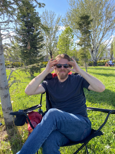 A man with a beard sits relaxed on a folding chair outdoors, wearing sunglasses and a dark t-shirt, surrounded by trees and lush green grass.