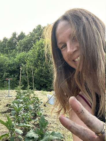 Une femme souriante aux cheveux longs prend un selfie dans un jardin, levant ses doigts boueux en signe de paix, entourée de plantes et d'arbres verdoyants.