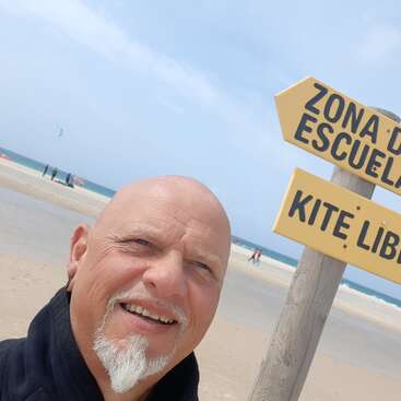 Ein lächelnder Mann mit Spitzbart macht am Strand ein Selfie. Hinter ihm weisen gelbe Schilder auf Spanisch auf ein Kiteboarding-Gebiet hin. Der Himmel ist klar.