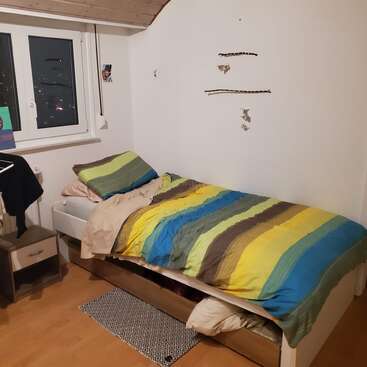 Una acogedora habitación con una cama individual con ropa de cama de rayas de colores, mesita de noche, ventana, decoraciones en la pared y cajones de almacenamiento debajo de la cama, todo ello sobre un suelo de madera.