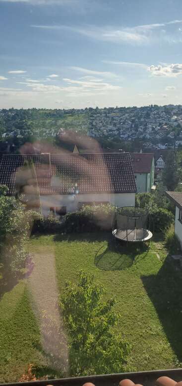 Una mano sosteniendo un teléfono se refleja en una ventana con vistas a un patio trasero con un trampolín, tejados, vegetación y una ciudad lejana bajo un cielo azul.