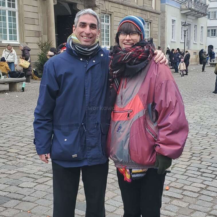 Un hombre y una mujer sonrientes posan juntos con ropa de invierno en una calle empedrada, con gente y edificios históricos visibles al fondo. Parece que hace frío.
