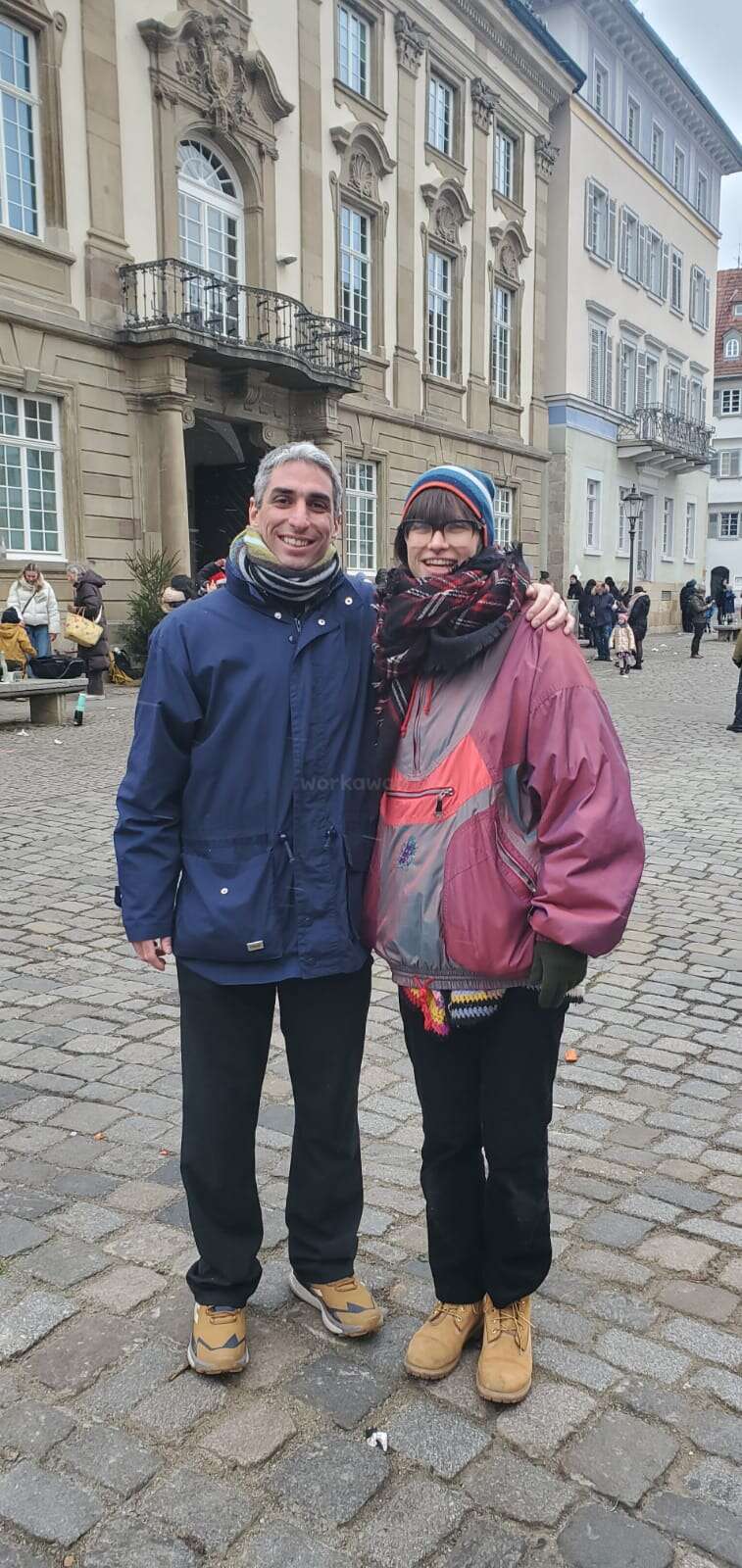 Un hombre y una mujer sonrientes posan juntos con ropa de invierno en una calle empedrada, con gente y edificios históricos visibles al fondo. Parece que hace frío.