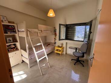Un acogedor dormitorio infantil con literas, una estantería con juguetes, una mesilla de noche amarilla, una silla de oficina, una ventana abierta y una suave luz natural.