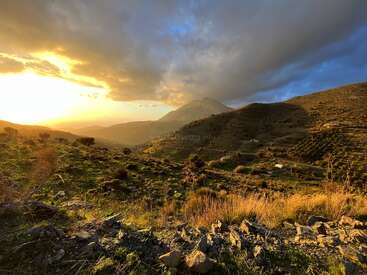 La luz dorada del sol baña una ladera rocosa y verde al atardecer. Dramáticas nubes se ciernen sobre las montañas distantes, creando un impresionante paisaje de calidez, sombra y belleza natural.