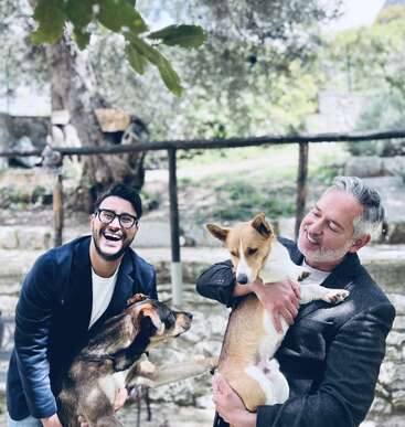 Dos hombres están al aire libre, sonriendo y riendo, mientras interactúan con dos perros. Un hombre sostiene a un perro cariñosamente, mientras el otro disfruta del momento juguetón y alegre.