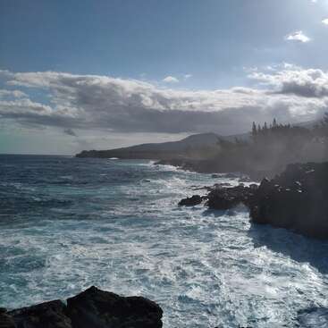 L'image représente un littoral accidenté avec des rochers sombres, des vagues écumantes et une chaîne de montagnes lointaine sous un ciel bleu avec des nuages blancs.