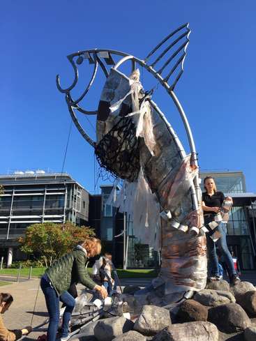 Das Bild zeigt eine große Metallskulptur eines Fisches, an der Menschen arbeiten, vor einem klaren blauen Himmel und einem modernen Gebäude im Hintergrund.