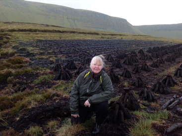 Das Bild zeigt eine Frau, die in einem Torffeld kniet, mit einer bergigen Landschaft im Hintergrund. Sie trägt eine grüne Jacke und eine schwarze Hose.