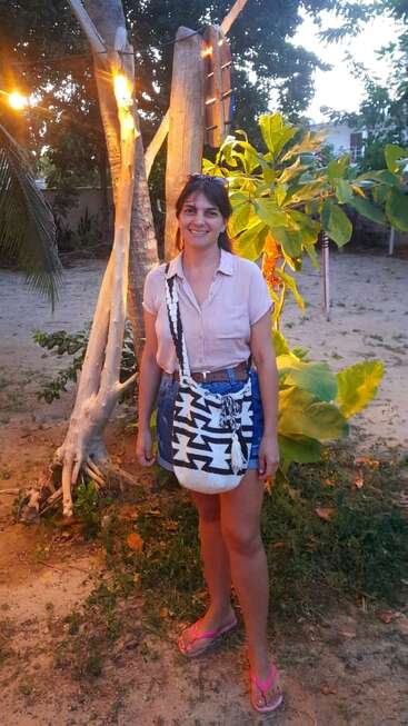 Eine Frau steht lächelnd im Freien und trägt eine leichte Bluse, einen Jeansrock, eine gemusterte Umhängetasche und Flip-Flops. Im Hintergrund sind Bäume, Lichterketten und sandiger Boden zu sehen. Das Abendlicht strahlt.
