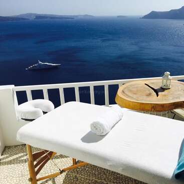 The image depicts a serene outdoor massage table setup on a balcony overlooking the ocean, with a white towel and a blue towel neatly rolled on top.