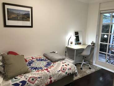 The image depicts a cozy bedroom with a bed, desk, and chair, featuring a vibrant comforter, framed picture, and French doors leading to an outdoor patio area.