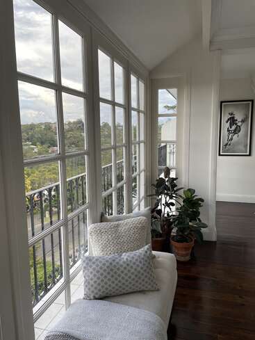 The image depicts a serene room with a large window offering a view of trees and houses, featuring a white couch, plants, and artwork, exuding a sense of calmness and tranquility.