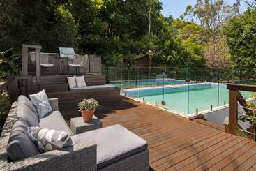 Esta imagem mostra um deck de madeira cheio de estilo com mesas ao ar livre, um vaso de plantas, uma área para churrasco e uma piscina cercada, rodeada por uma vegetação exuberante.