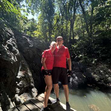 Un couple souriant, vêtu de chemises rouges assorties, se tient sur un pont en bois au bord d'un étang naturel, entouré d'une forêt verdoyante et d'un terrain rocailleux, sous l'effet de la lumière du soleil.