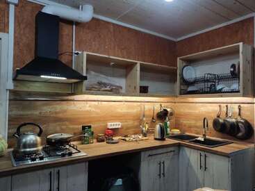 This cozy kitchen features wooden cabinets, open shelving, a black stove hood, hanging utensils, dish rack, kettle, pots, pans, and a gas stove setup.