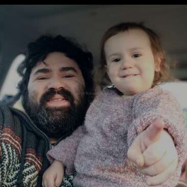 Das Bild zeigt einen Mann mit dunklen Haaren und Bart, der einen gemusterten Pullover trägt und mit einem Kleinkind in einem rosa Pullover zusammensitzt, das auf die Kamera zeigt.