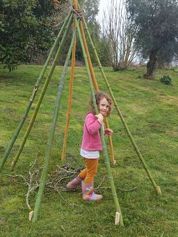 Une jeune fille chaussée de bottes colorées se tient debout à l'intérieur d'une structure en bambou en forme de tipi, posée sur l'herbe et entourée de branches, dans un cadre naturel en plein air avec des arbres en arrière-plan.