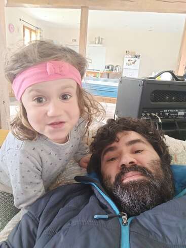 Un homme barbu est allongé, souriant, tandis qu'une petite fille aux cheveux bouclés, coiffée d'un bandeau rose, prend la pose à ses côtés dans une pièce chaleureuse.