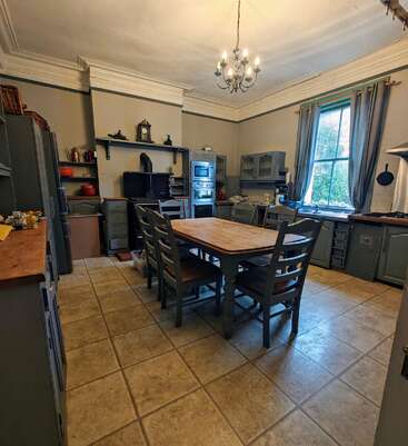 This image shows a spacious, traditional kitchen with tile flooring, a wooden dining table, vintage cabinets, a window with curtains, and a decorative chandelier overhead.