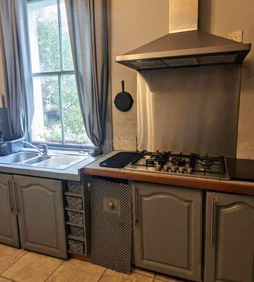 This kitchen features a gas stove, metal extractor hood, blue-gray cabinets, a sink by a window with gray curtains, and a hanging cast iron skillet.