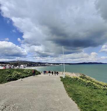 A group of people walk along a seaside pathway under a dramatic sky, with green grass, distant town buildings, and calm blue-green sea beside them.