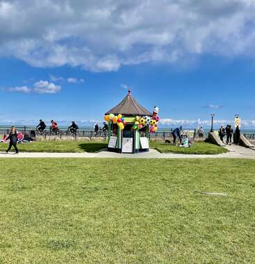 A small gazebo decorated with colorful balloons stands near the seaside. People walk and cycle along the promenade under a blue sky with scattered clouds.