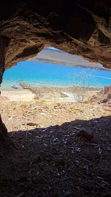A imagem mostra uma vista incrível de dentro de uma caverna rochosa, com vista para um lago turquesa, vegetação seca, montanhas distantes e um pequeno barco atracado na margem.