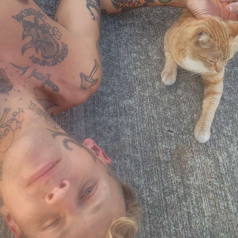 L'image représente un homme allongé sur le sol avec un chat, mettant en évidence ses tatouages et le pelage orange du chat aux pattes blanches. Le visage de l'homme est flou.