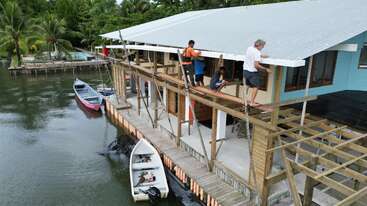 Four people are working on the balcony construction of a waterfront house. Several boats are docked nearby, surrounded by lush green palm trees and calm water.