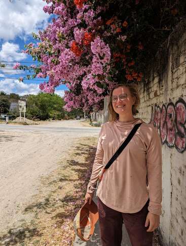 Eine lächelnde Person steht an einer mit Graffiti bedeckten Wand, unter leuchtend rosa und orangefarbenen Blumen, mit einem Hut in der Hand, in helles Tageslicht getaucht. Darüber ein blauer Himmel.