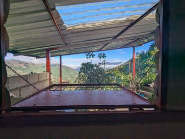 Der Blick durch ein Fenster zeigt sattgrüne Hügel, einen blauen Himmel mit Wolken, ein Metalldach und eine hölzerne Plattform, die eine ruhige Bergatmosphäre schaffen.