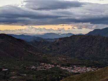 Ein kleines Dorf schmiegt sich in ein üppig grünes Tal, umgeben von dramatischen Bergen, unter einem stimmungsvollen Himmel mit Wolkenschichten und verblassendem Abendsonnenlicht.