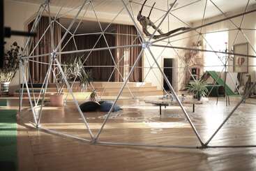 Une pièce spacieuse et lumineuse, dotée d'une grande structure en dôme géodésique, de plantes d'intérieur, de coussins, d'une petite table, d'une scène avec des rideaux et de motifs artistiques au sol partout dans la pièce.