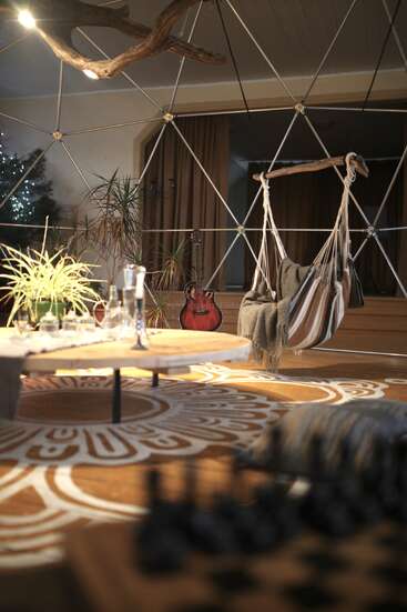 Un espace de vie chaleureux et moderne, agrémenté d'un fauteuil suspendu, de plantes, d'une guitare, d'une structure géométrique en forme de dôme, d'une table ronde en bois, d'un tapis à motifs et d'un échiquier, le tout baigné d'une lumière tamisée.