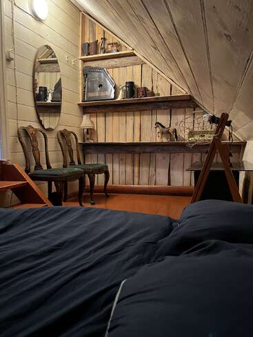 Cette chambre mansardée au charme douillet se distingue par ses murs en bois rustiques, ses chaises vintage, ses étagères garnies d'antiquités, son miroir ovale, sa literie aux tons sombres et son éclairage tamisé et chaleureux, qui créent une atmosphère nostalgique.