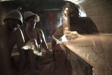 Quatre personnes sont assises ensemble dans un sauna en bois, vêtues de vêtements légers et coiffées de chapeaux en feutre. L'atmosphère est embuée, intime et détendue, baignée d'une lumière jaune et chaleureuse.