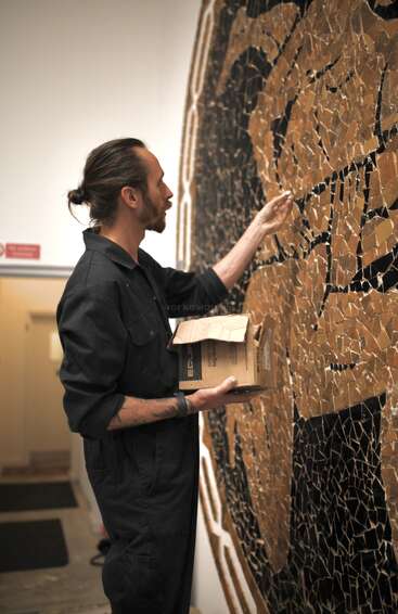 Un homme vêtu d'une combinaison sombre pose avec soin des carreaux sur une grande mosaïque. Il tient une boîte dans les mains, concentré sur la réalisation de ce motif complexe aux teintes brun doré.