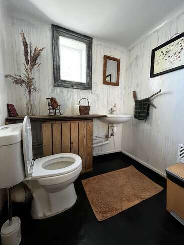Cette salle de bains au style rustique comprend des toilettes blanches, un petit lavabo, des plantes séchées décoratives, des touches de couleur terre, des décorations murales, une fenêtre encadrée, des miroirs et un tapis de sol marron.