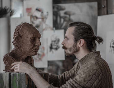 Un artiste concentré, moustachu et les cheveux attachés en arrière, examine un buste en argile, entouré de diverses esquisses artistiques épinglées sur le mur de l'atelier derrière lui.