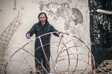 Un homme vêtu d'une combinaison sombre se tient debout dans un espace industriel délabré, occupé à travailler sur de grandes sculptures géométriques réalisées en fil de fer. En arrière-plan, on aperçoit de la peinture écaillée.