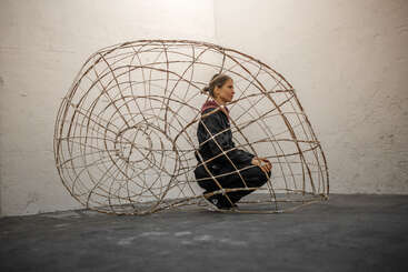 Une femme vêtue de noir est accroupie à l'intérieur d'une grande structure métallique en spirale qui ressemble à un coquillage, dans une pièce dépouillée et minimaliste au sol gris.