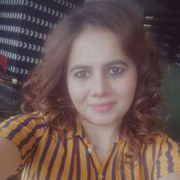 A woman with curly brown hair smiles at the camera, wearing a yellow and navy striped shirt. The background has patterned walls and a cozy ambiance.