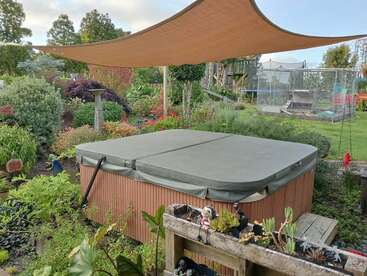 Ein überdachter Whirlpool steht in einem üppigen, bunten Garten unter einem großen Sonnensegel. Im Hintergrund gibt's ein Trampolin und ein Holzspielgerät.