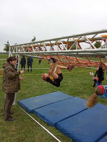 Children wearing red headbands navigate a rope obstacle course, hanging upside down. An adult supervises nearby. Mats are placed below for safety on the grassy field.