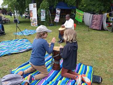 Two children kneel on striped blankets, drumming together outdoors. A man in white plays a drum nearby under a tent, with colorful banners and posters behind him.