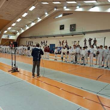 Un groupe d'escrimeurs en uniforme blanc s'aligne à l'intérieur d'un gymnase. Un officiel semble s'adresser à eux. Le décor est planté pour une compétition.