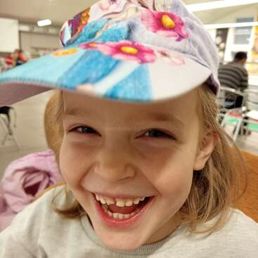 A joyful child with long hair laughs brightly, wearing a colorful cap with floral designs. The indoor setting is lively, with people and tables in the background.